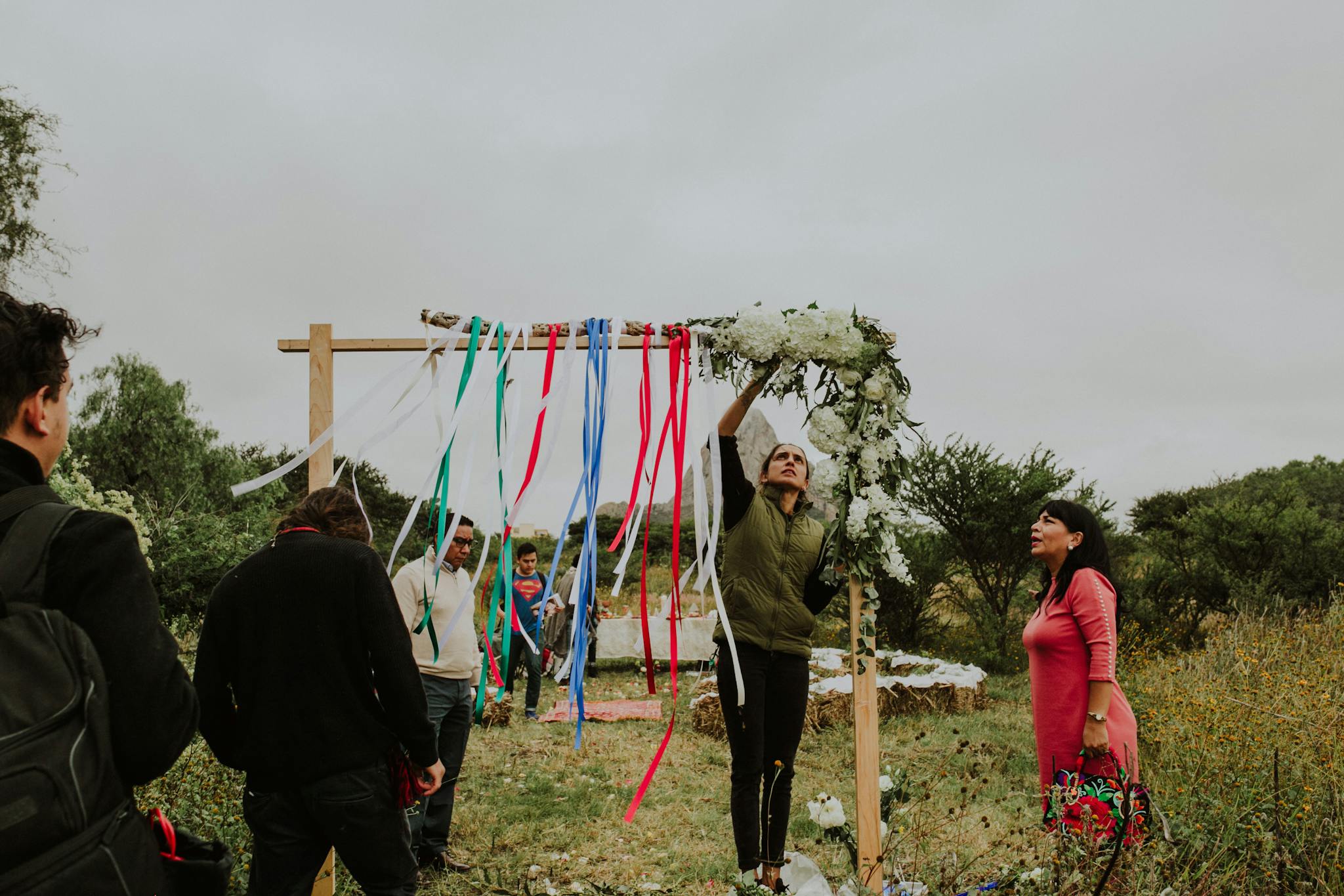 People preparing a colorful outdoor wedding arch with ribbons and flowers in a natural setting.